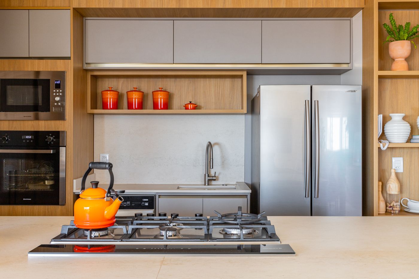 Cozinha clara com marcenaria amadeirada, nichos abertos com potes vermelhos, cooktop com chaleira laranja e geladeira inox