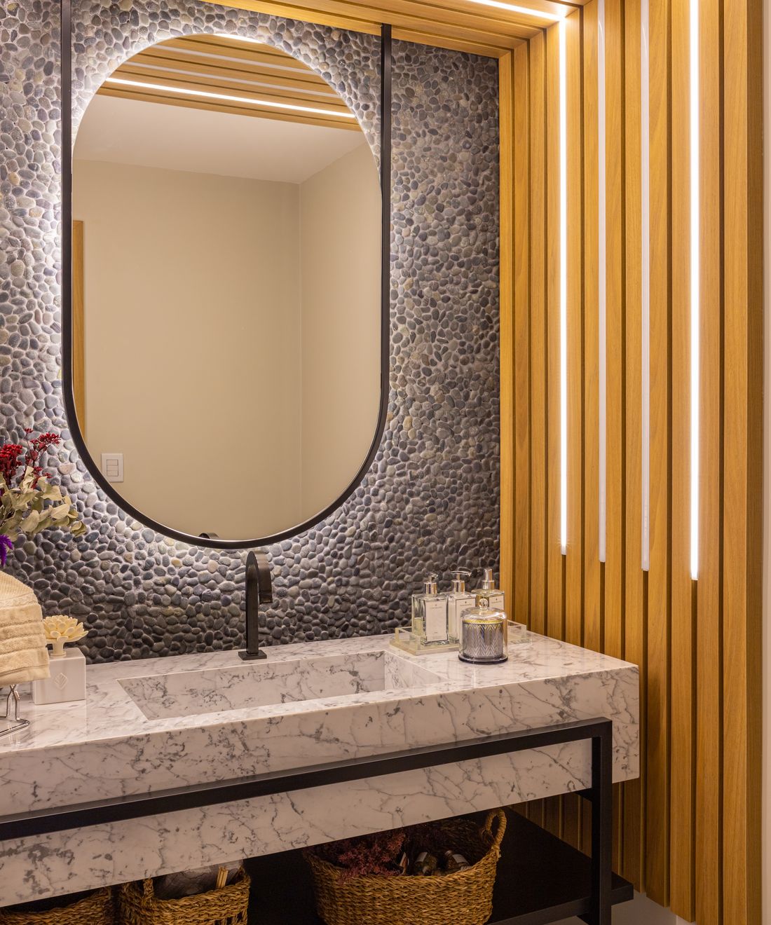 Lavabo com painel ripado de madeira, espelho oval, parede de seixos e bancada de mármore sobre estrutura metálica preta