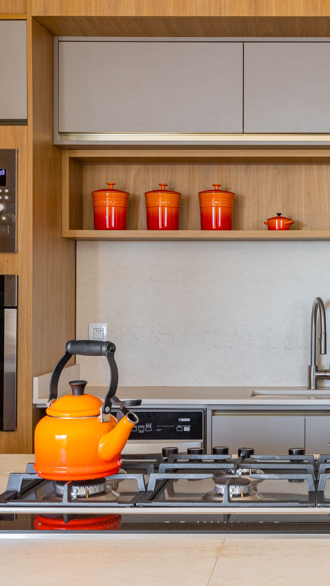 Cozinha com marcenaria amadeirada, nicho aberto com potes vermelhos e cooktop com chaleira laranja