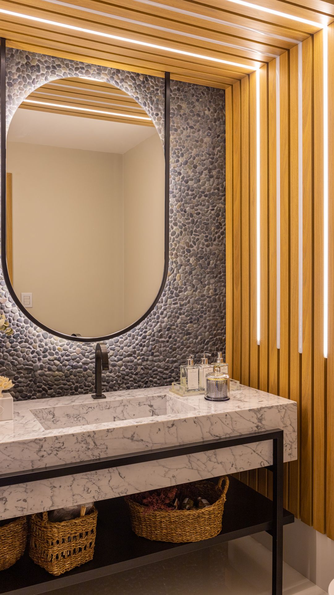 Lavabo com painel ripado de madeira, espelho oval, parede de seixos e bancada de mármore sobre estrutura metálica preta
