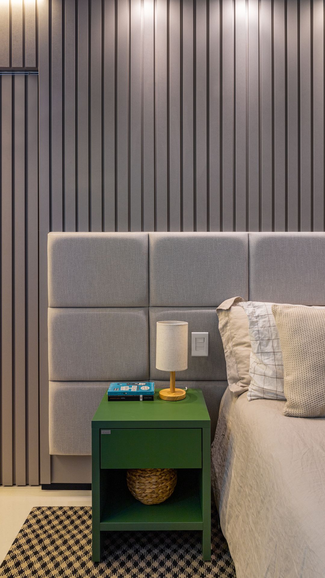 Quarto com cabeceira estofada em módulos, painel ripado chumbo e criado-mudo verde com abajur
