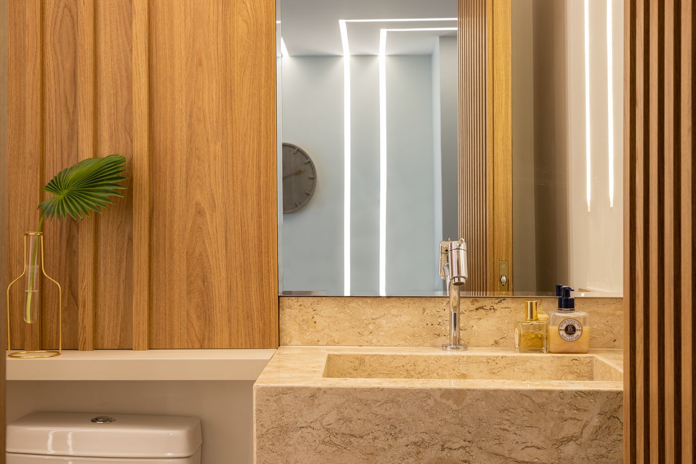 Lavabo com cuba esculpida em pedra, espelho com frisos de LED e painel ripado de madeira