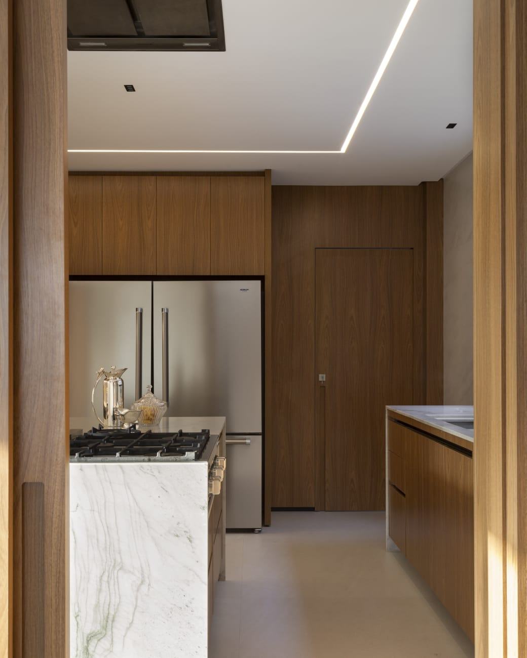Cozinha em madeira nogueira com porta invisível, geladeira embutida e ilha com cooktop e mármore em cascata