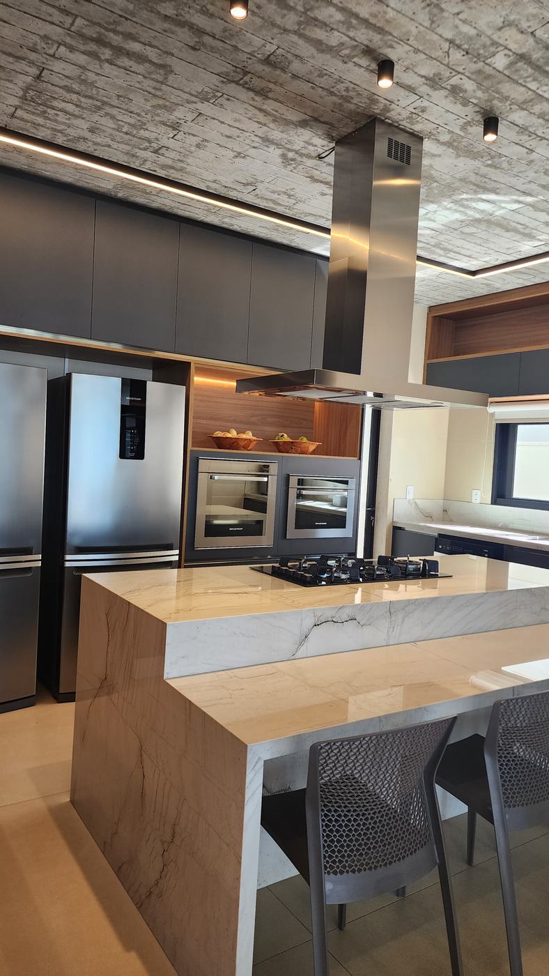 Cozinha grafite com torre de fornos, nicho amadeirado iluminado e ilha em quartzito com cooktop e banquetas