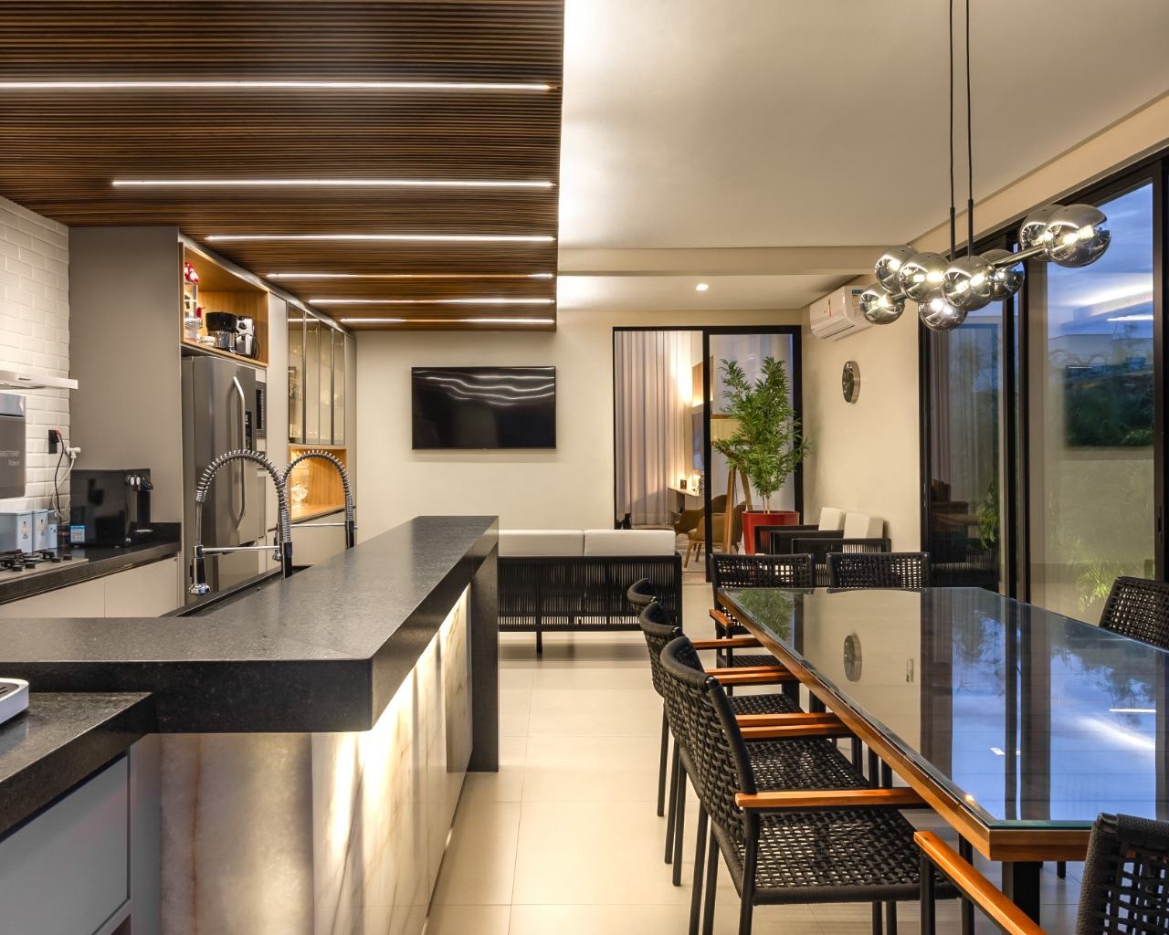 Espaço gourmet com bancada de granito preto, frontão de ônix iluminado, marcenaria cinza com nichos amadeirados e forro ripado