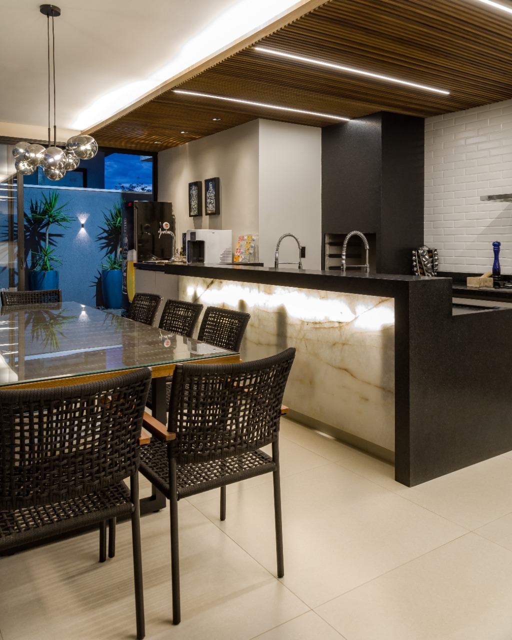 Espaço gourmet com bancada e churrasqueira em granito preto, frontão de ônix iluminado e forro ripado de madeira com LED