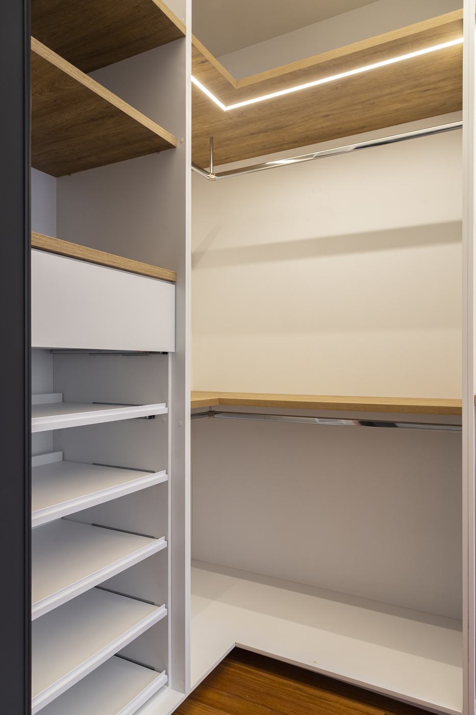 Closet branco em L com prateleiras amadeiradas, fita de LED, cabideiros cromados e sapateira de prateleiras deslizantes