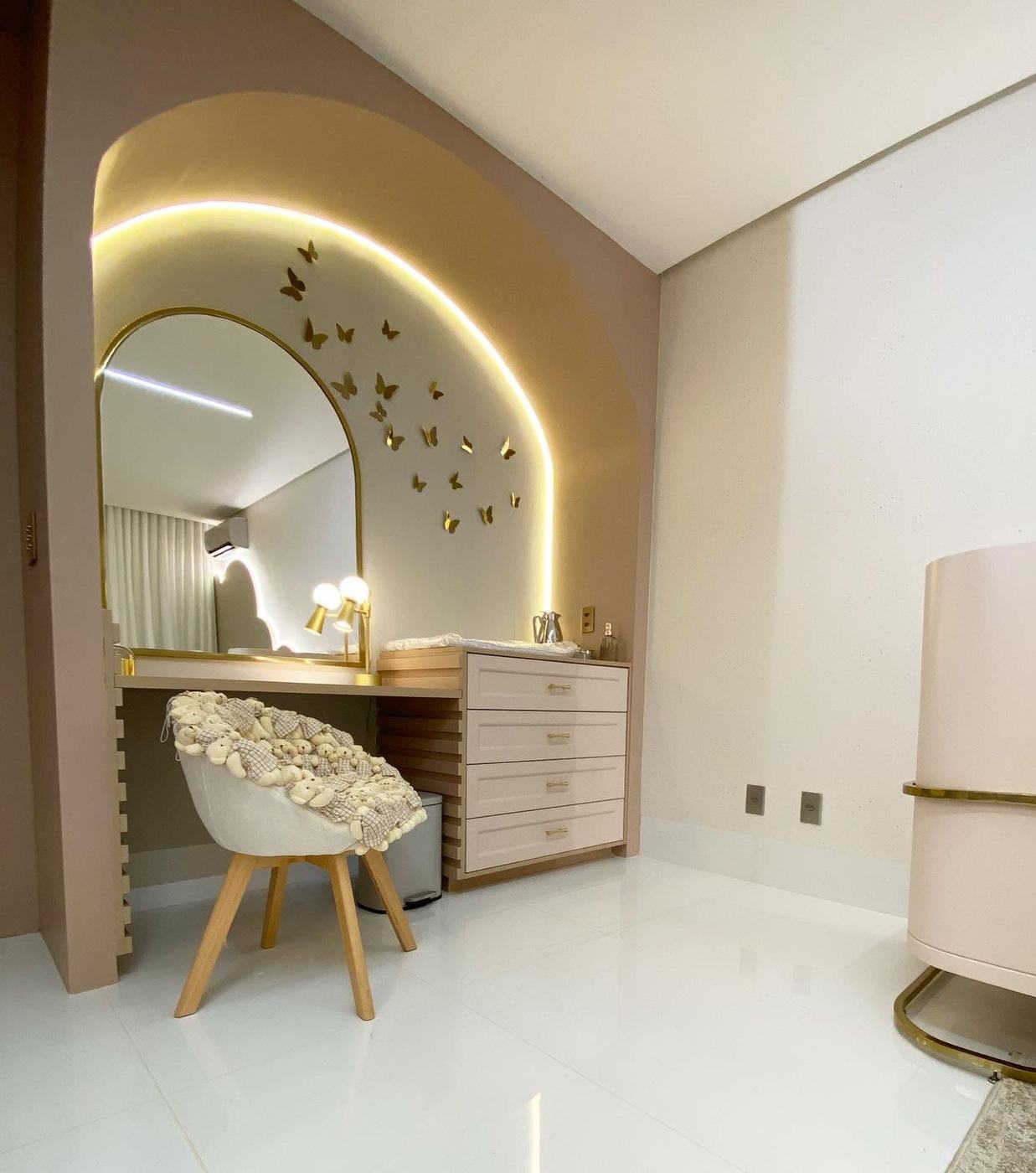 Quarto de bebê com bancada e cômoda trocador em laca rosé, puxadores dourados, base ripada e nicho em arco iluminado