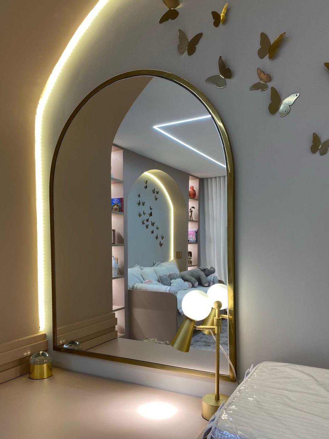 Espelho em arco com moldura dourada sobre a bancada rosé, refletindo a cama-baú com estantes iluminadas